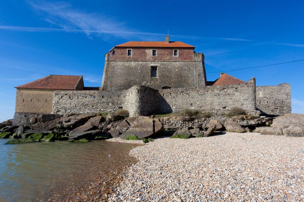 Une forteresse historique en pierre avec un toit de tuiles rouges située sur une plage de galets contre un ciel bleu clair.