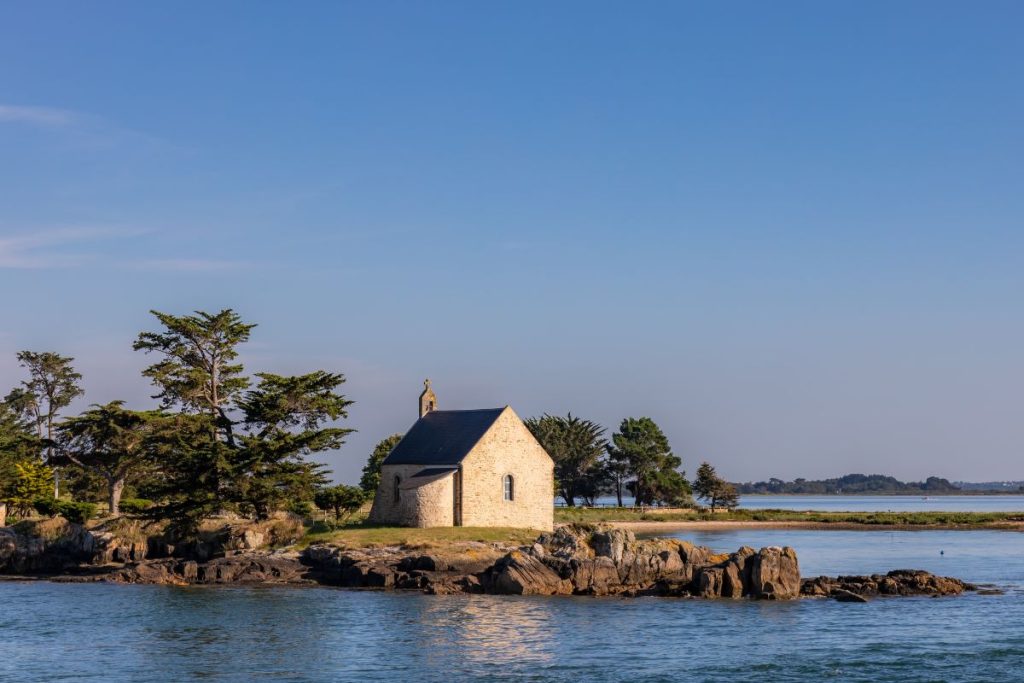 Chapelle en pierre isolée sur une petite île entourée d'eau, avec arbres et ciel bleu en arrière-plan.
