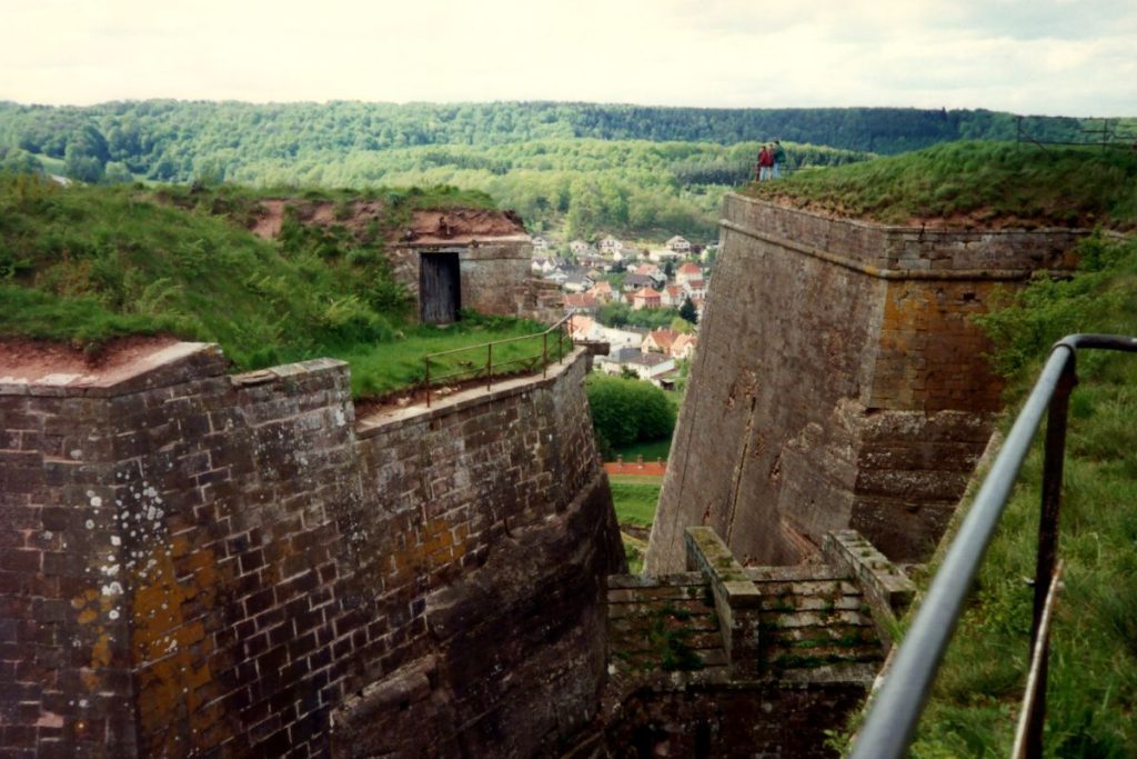 Une personne se tient au sommet d’un ancien mur de forteresse surplombant une petite ville européenne nichée dans une vallée verdoyante et luxuriante.