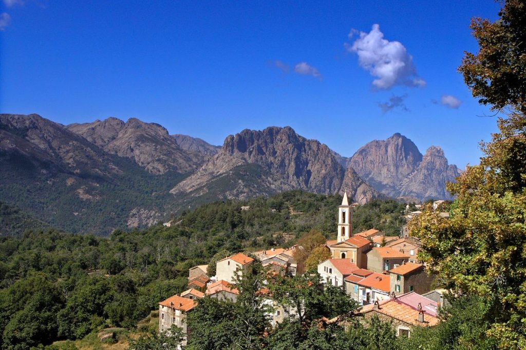 Une vue panoramique sur un village de montagne avec un clocher d'église proéminent sous un ciel bleu avec des nuages clairsemés.