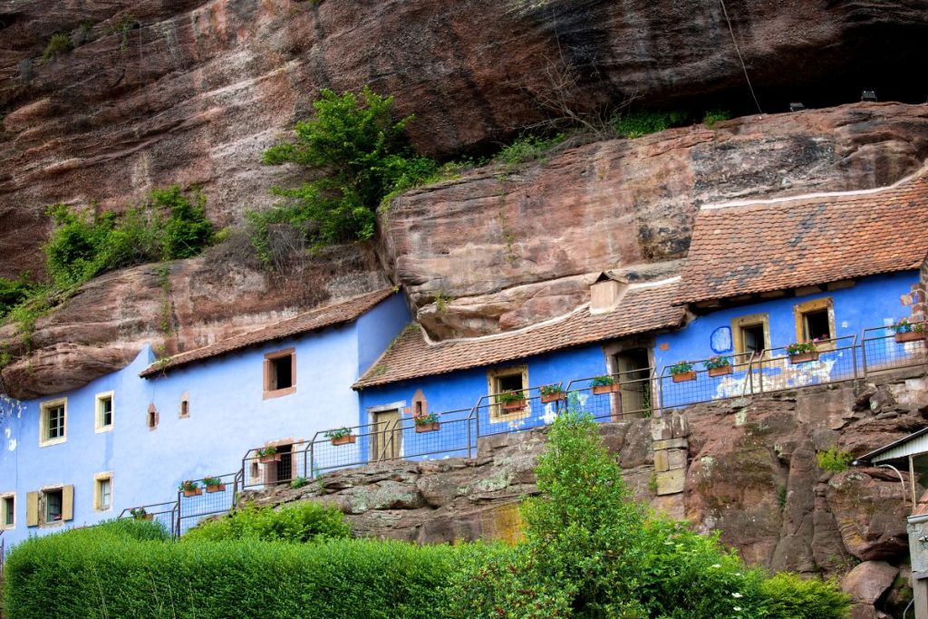Une maison bleue construite sur le flanc d’une falaise rocheuse avec de la végétation au-dessus.
