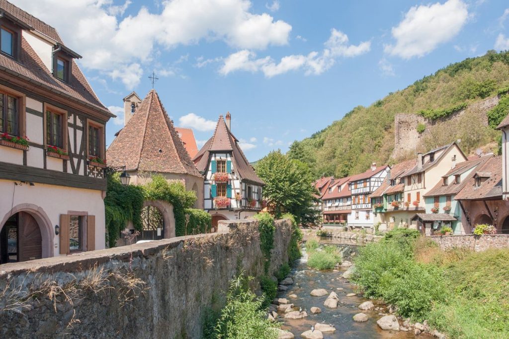 Village européen pittoresque avec des maisons traditionnelles à colombages au bord d'une petite rivière, sous un ciel bleu clair.