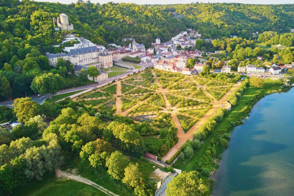 Vue aérienne d'un jardin à la française au bord d'une rivière, avec un complexe de bâtiments historiques et des collines verdoyantes en arrière-plan.