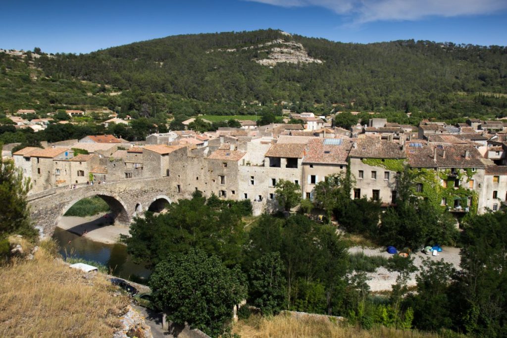 Une vue panoramique sur un village européen traditionnel avec des maisons en pierre, un vieux pont traversant une rivière, entouré de verdure luxuriante et de collines.