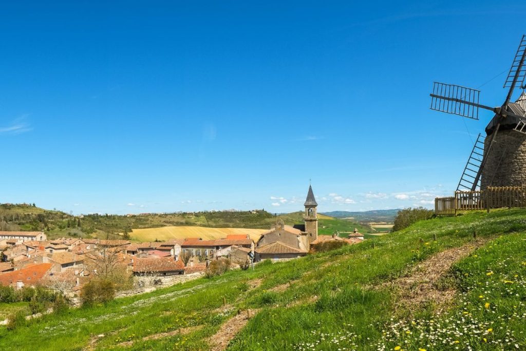 Paysage rural avec un moulin à vent traditionnel surplombant un village et des terres agricoles sous un ciel bleu clair.