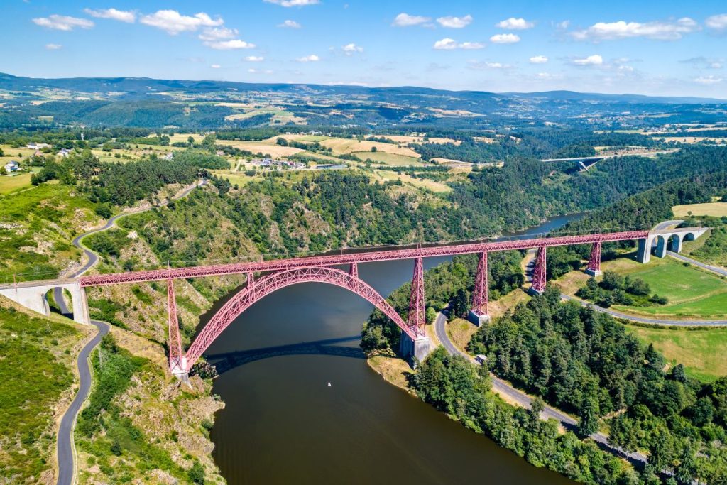 Vue aérienne d'un pont voûté rouge enjambant une rivière dans un paysage vallonné et luxuriant.