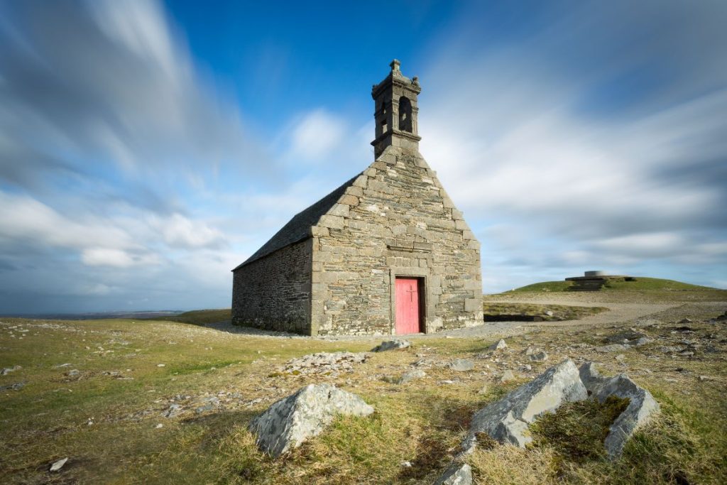 Chapelle en pierre avec porte rouge sous un ciel dynamique sur un terrain herbeux et rocheux.