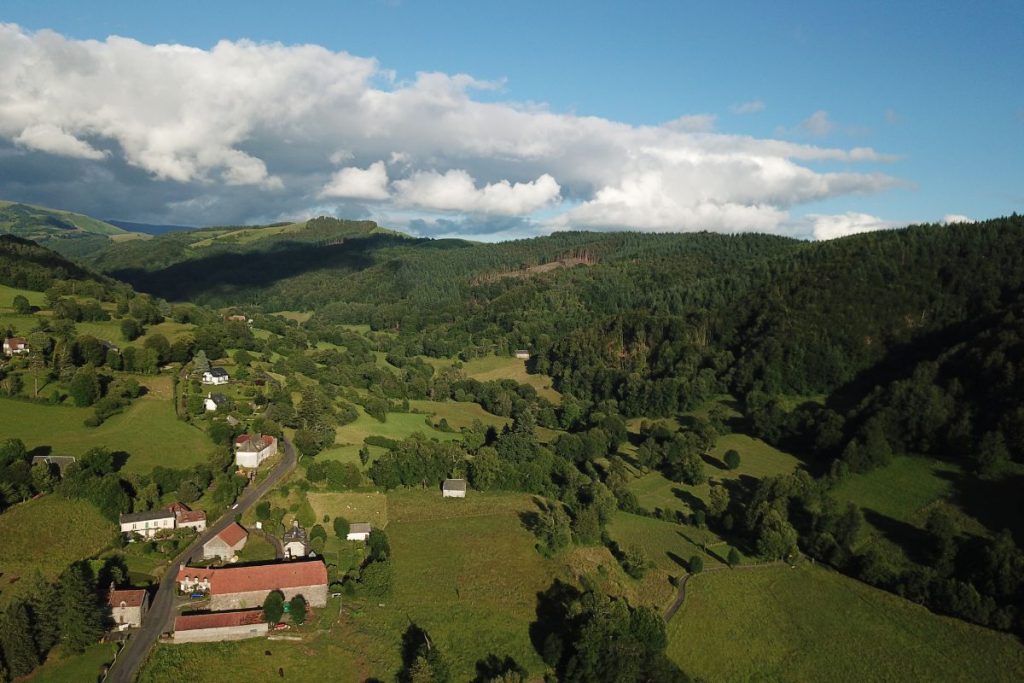 Vue aérienne d'un paysage rural avec une verdure luxuriante et des maisons dispersées sous un ciel partiellement nuageux.