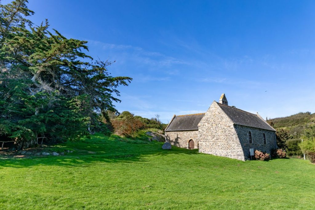 Une petite chapelle en pierre au toit incliné entourée d'herbe verte sous un ciel bleu clair avec des arbres épars.