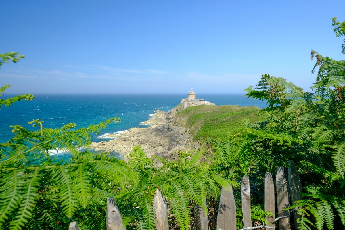 Un phare situé sur un promontoire côtier, entouré de verdure sous un ciel bleu clair.