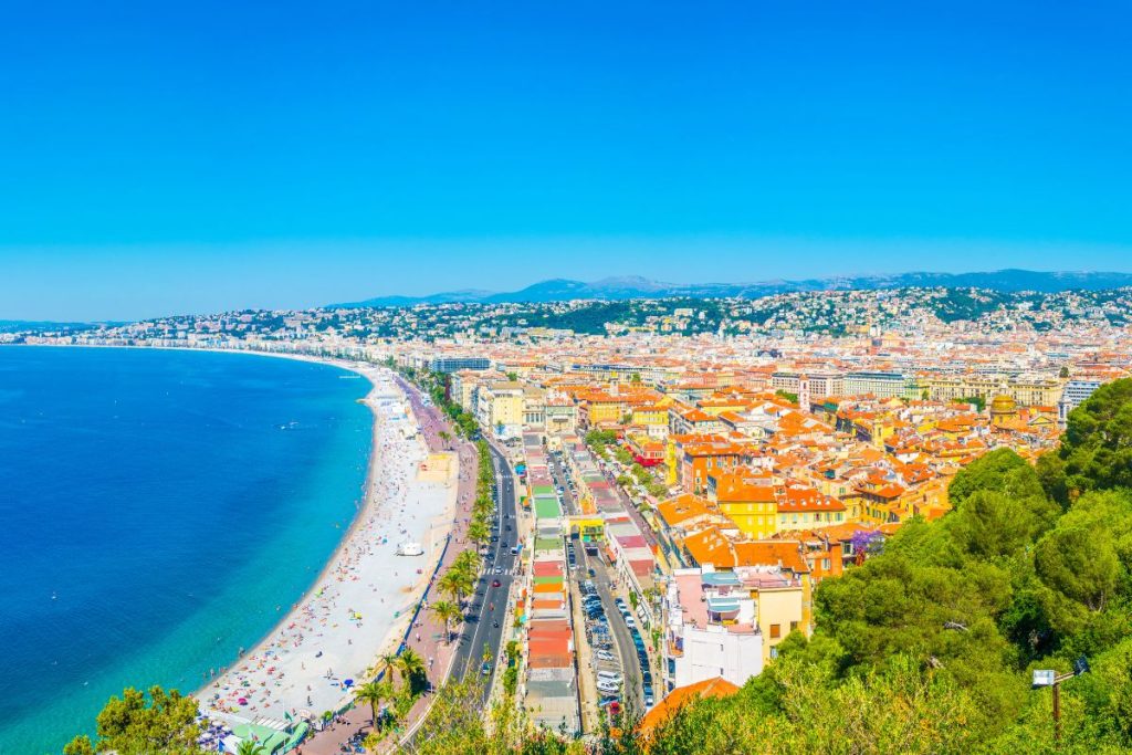 Vue panoramique d'une ville côtière animée avec un front de mer, des eaux bleues et des bâtiments colorés sous un ciel clair.
