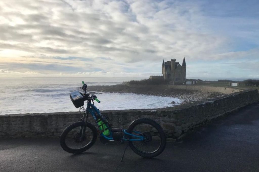 Un vélo garé sur un chemin en bord de mer avec vue sur un vieux château et la mer en arrière-plan.
