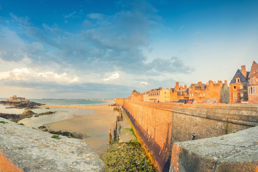 Ville côtière historique avec murs en pierre surplombant une plage de sable à marée basse.
