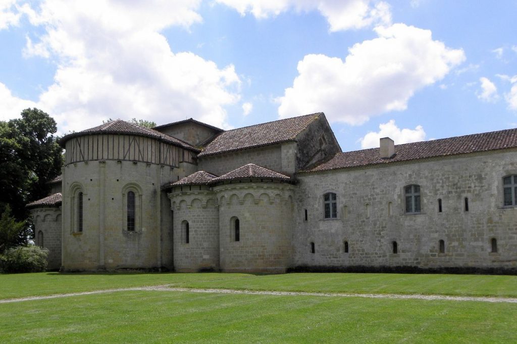 Ancienne église en pierre avec absides semi-circulaires et sections rectangulaires, entourée d'une pelouse verte sous un ciel généralement dégagé.