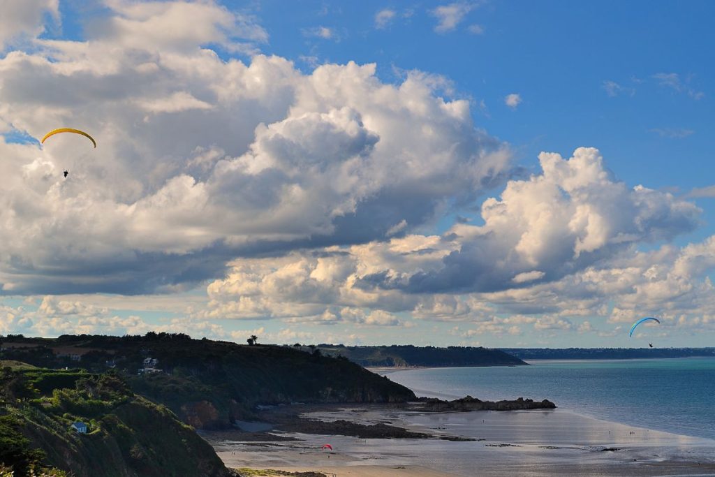 Parapentes survolant un paysage côtier avec des falaises et une plage sous un ciel bleu nuageux.