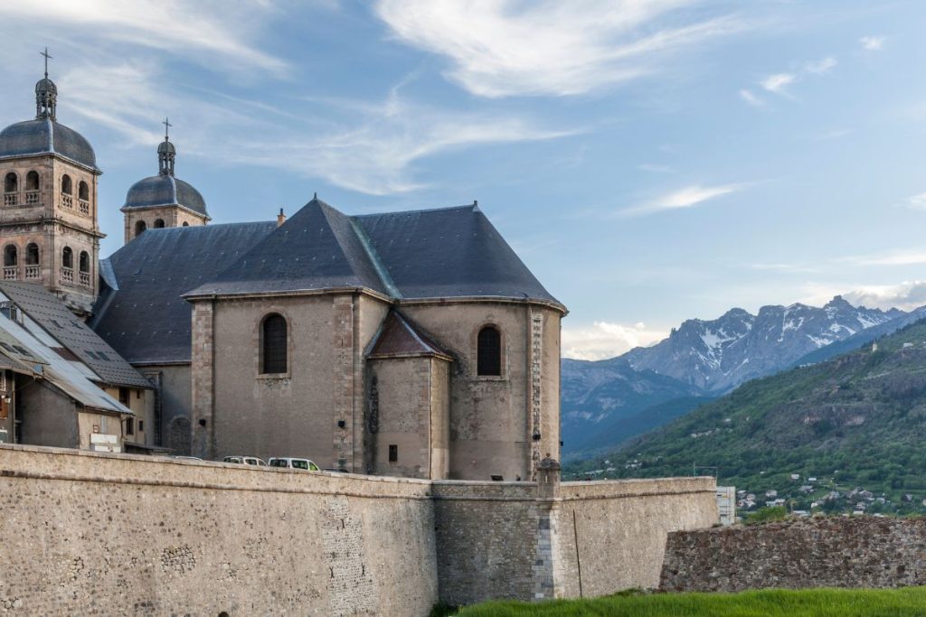 Église historique en pierre avec un mur fortifié au premier plan, sur fond de collines verdoyantes et de lointaines montagnes enneigées sous un ciel clair.