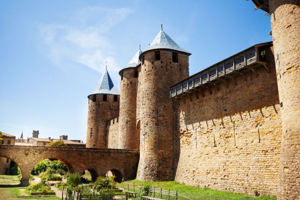 Un château médiéval en pierre avec deux grandes tours et un pont en bois sous un ciel bleu clair.