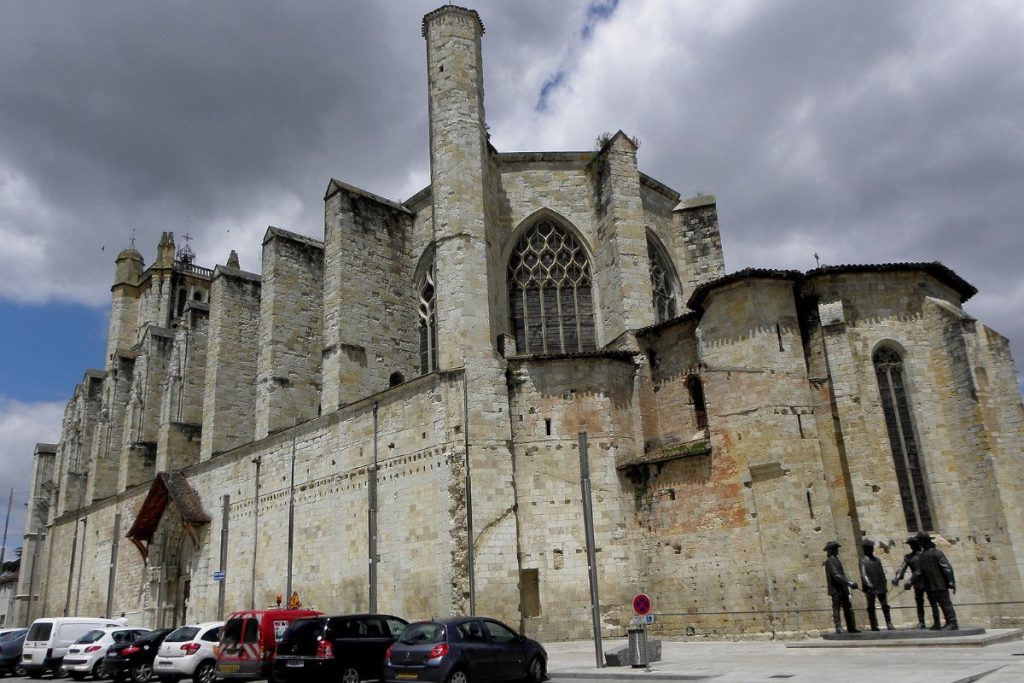 Vue extérieure d'une vieille cathédrale gothique avec de grands murs en pierre et des vitraux, sous un ciel nuageux, avec plusieurs personnes marchant à proximité.
