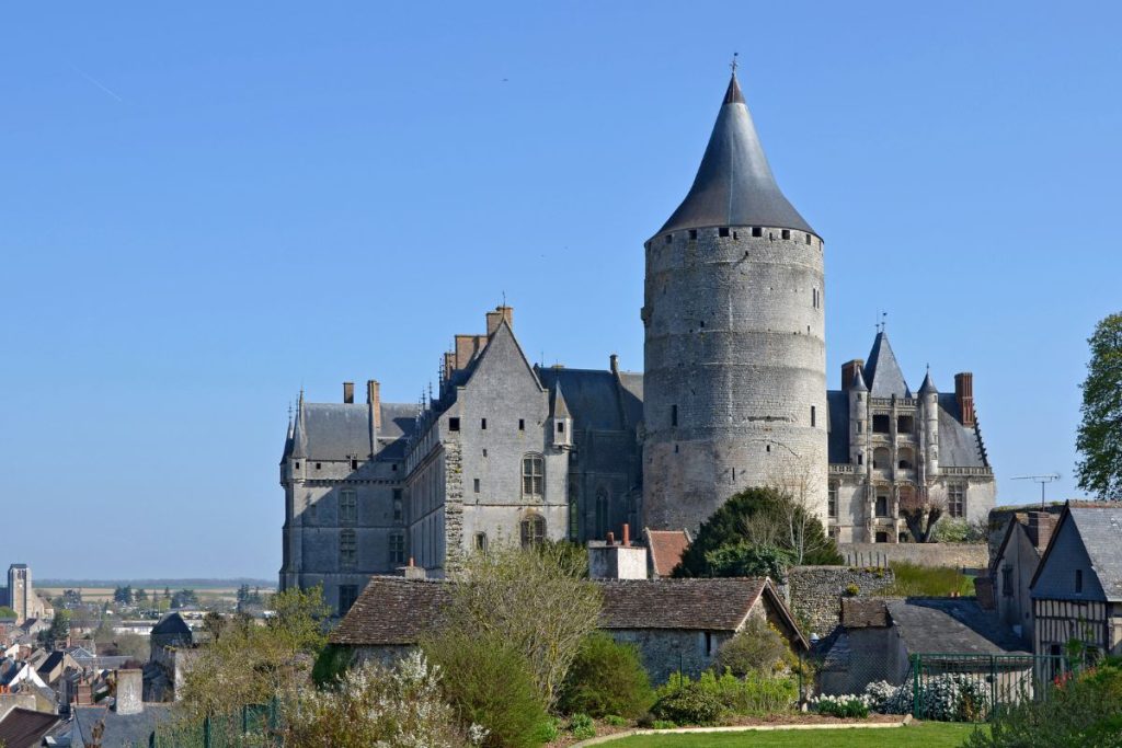 Château médiéval avec une tour ronde proéminente et des bâtiments attenants, sur fond de ciel bleu clair, surplombant un petit village européen.