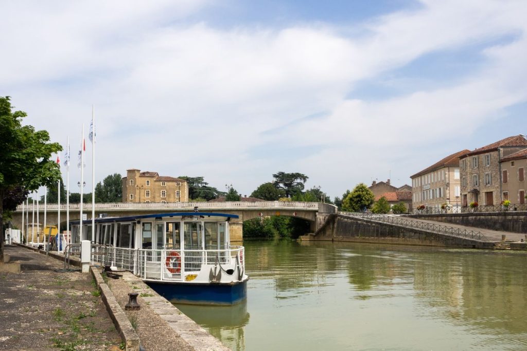 Un bateau d'excursion amarré sur une rivière calme à côté d'une passerelle, avec un pont et des bâtiments historiques en arrière-plan sous un ciel clair.
