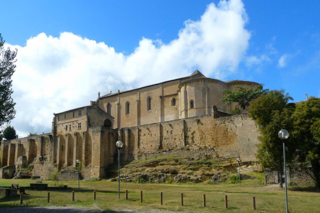 Ancienne abbaye fortifiée aux contreforts proéminents sous un ciel bleu avec des nuages.