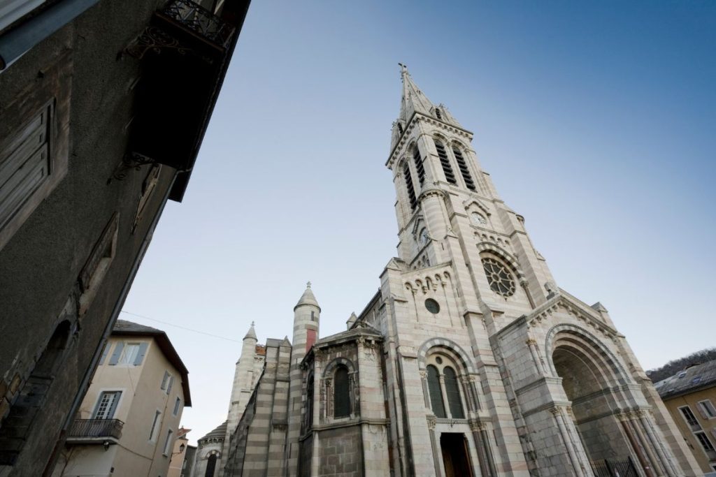 Une église de style gothique avec une haute flèche, encadrée par les bâtiments environnants sous un ciel dégagé.