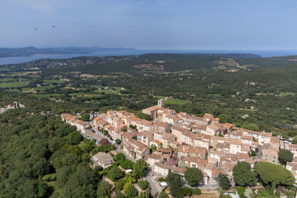 Vue aérienne d'un village historique avec des bâtiments en pierre et une église importante, entouré d'une campagne luxuriante et d'un littoral lointain.
