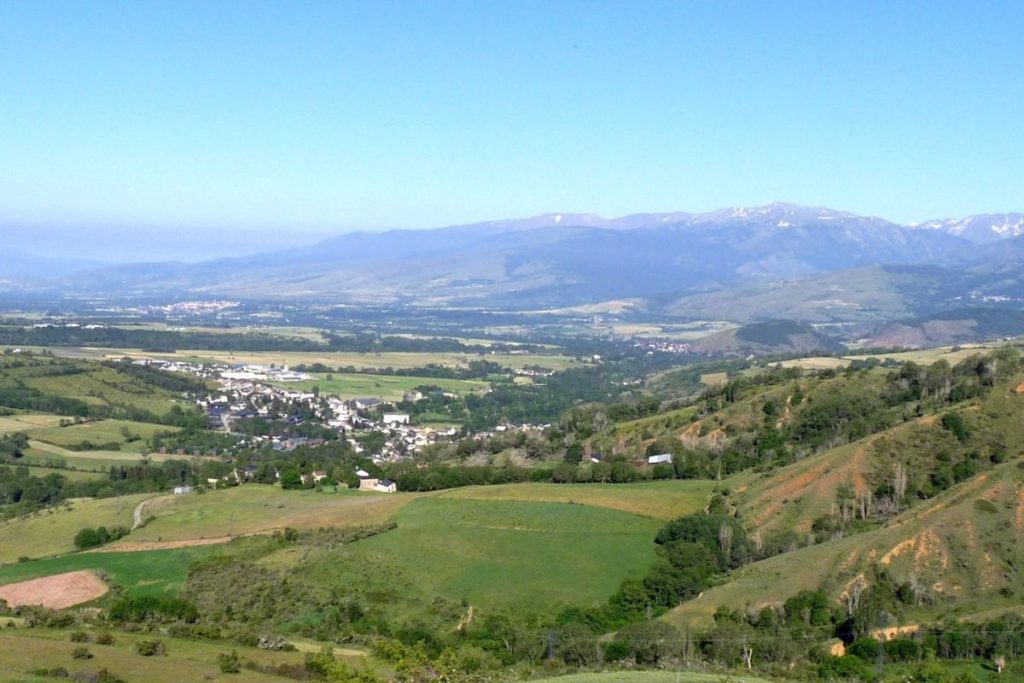 Vue panoramique sur une vallée luxuriante avec une petite ville, des champs verdoyants et des montagnes lointaines sous un ciel bleu clair.