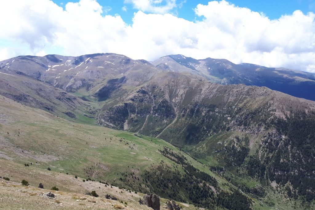 Vue panoramique sur une chaîne de montagnes avec des pentes verdoyantes et des plaques de neige sous un ciel nuageux.