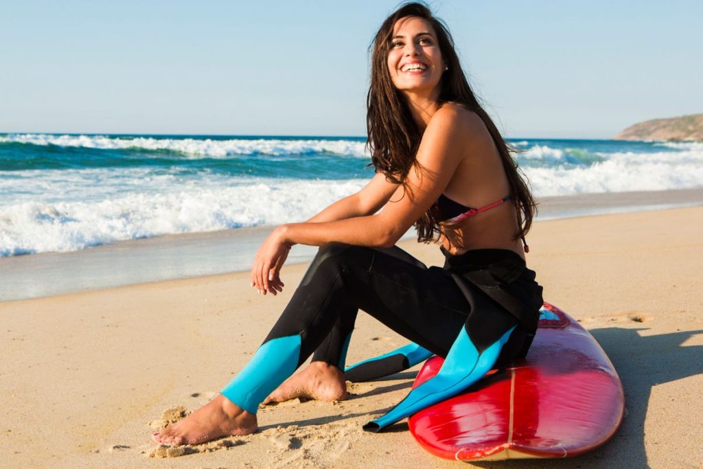 Une femme souriante en combinaison est assise sur une planche de surf rouge sur la plage, avec des vagues et un ciel clair en arrière-plan.