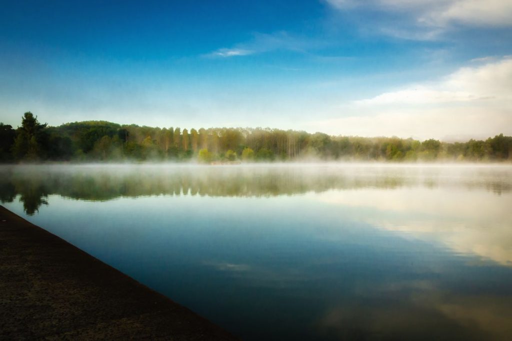 Brume matinale sur un lac serein avec des arbres luxuriants se reflétant dans l'eau calme, sous un ciel bleu.