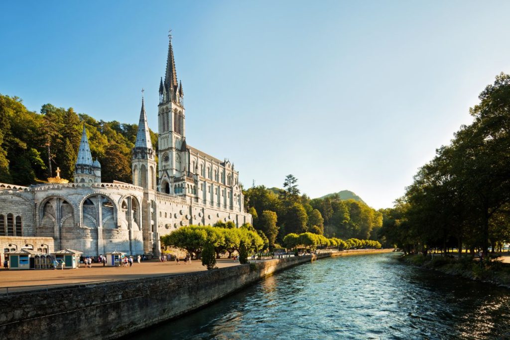 Le sanctuaire de Notre-Dame de Lourdes au bord d'une rivière, présentant une architecture richement ornée de flèches, dans un paysage serein bordé d'arbres pendant la journée.