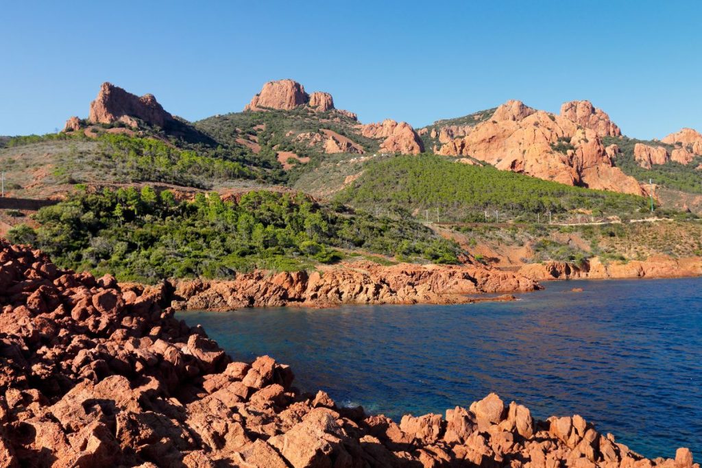 Vue panoramique sur un littoral accidenté avec des formations de roches rouges et une verdure luxuriante sous un ciel bleu clair.