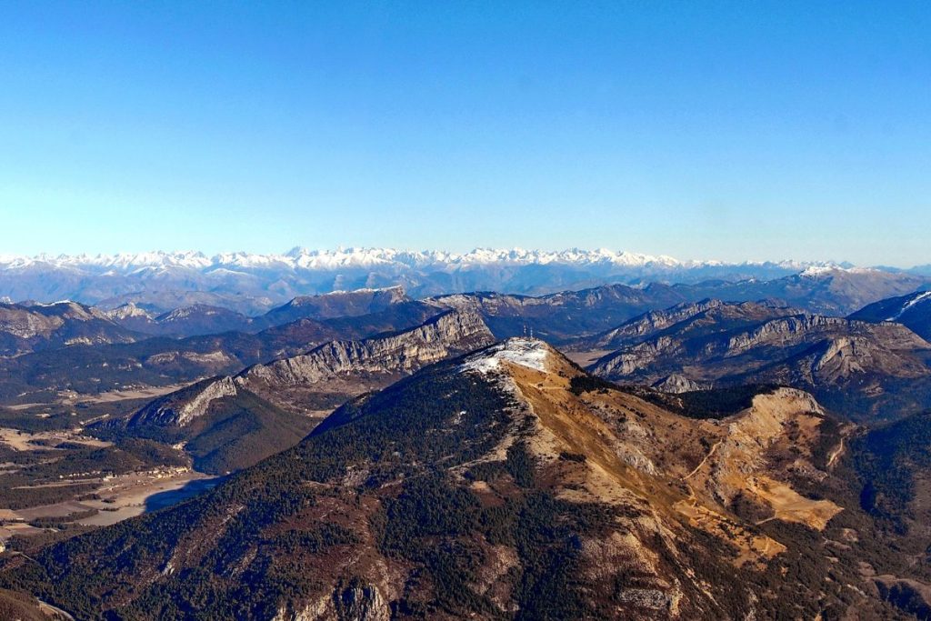 Vue aérienne d'un terrain montagneux avec des sommets enneigés au loin sous un ciel bleu clair.