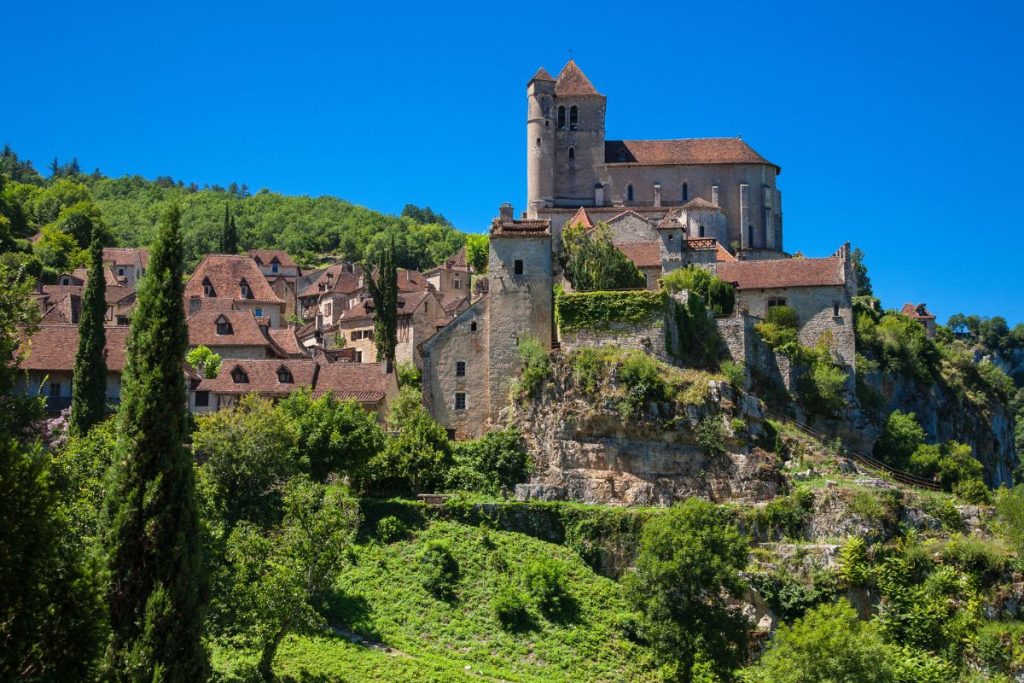 Une vue panoramique sur un village perché avec une église en pierre, niché au milieu d'une verdure luxuriante sous un ciel bleu clair.