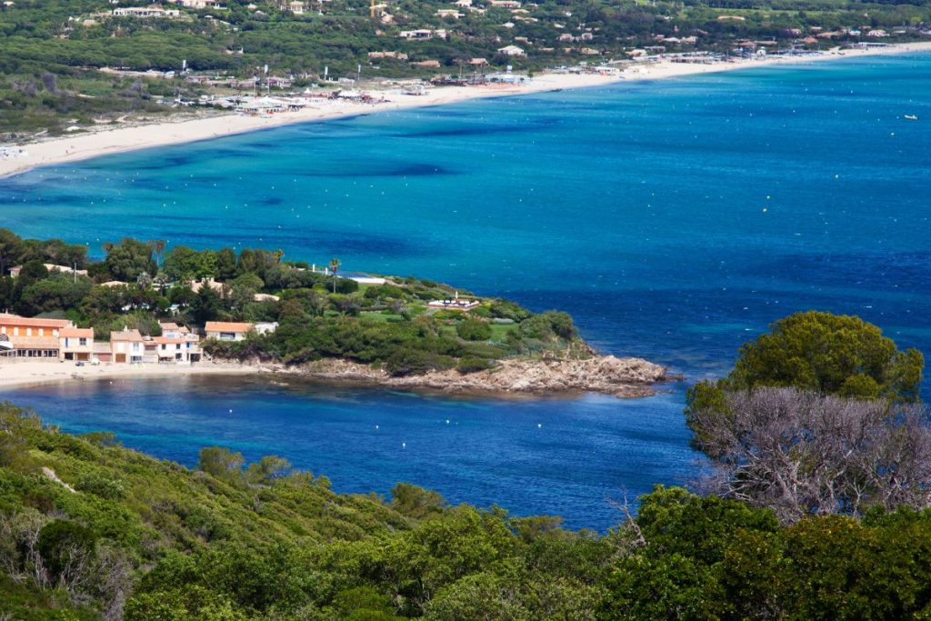 Vue aérienne d'une zone côtière présentant des eaux bleu clair, des plages de sable fin et un feuillage vert avec des bâtiments dispersés.