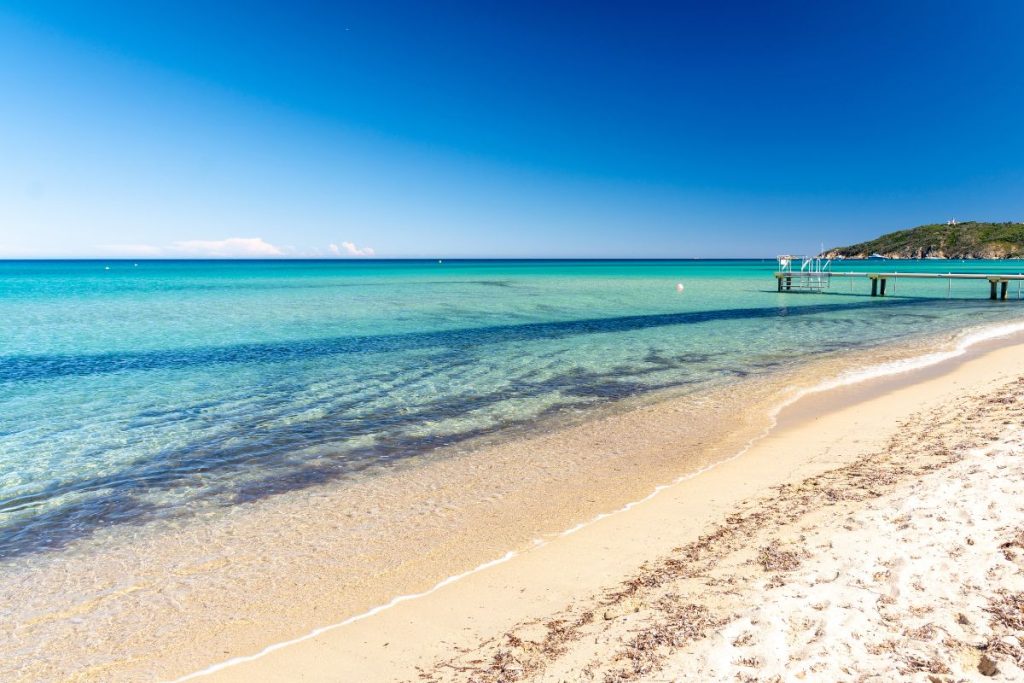 Une plage immaculée aux eaux turquoise, du sable blanc, un petit quai au loin, sous un ciel bleu éclatant.
