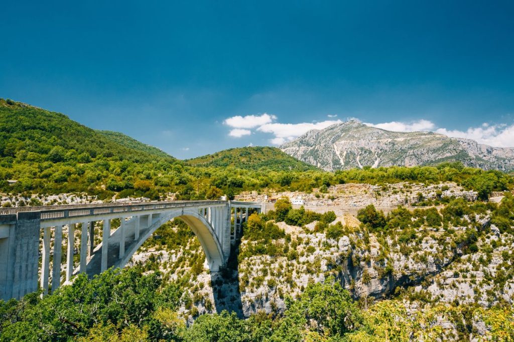 Pont en arc enjambant un canyon avec une verdure luxuriante et des montagnes rocheuses sous un ciel bleu clair.