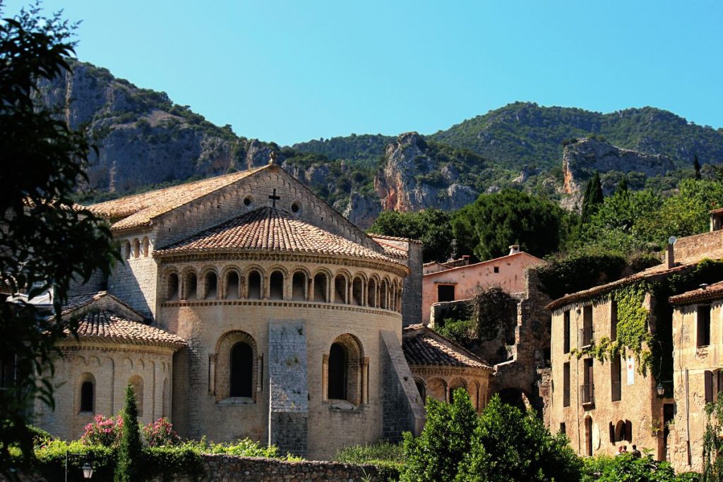 Ancienne église en pierre et bâtiments environnants nichés dans un paysage montagneux verdoyant sous un ciel bleu clair.