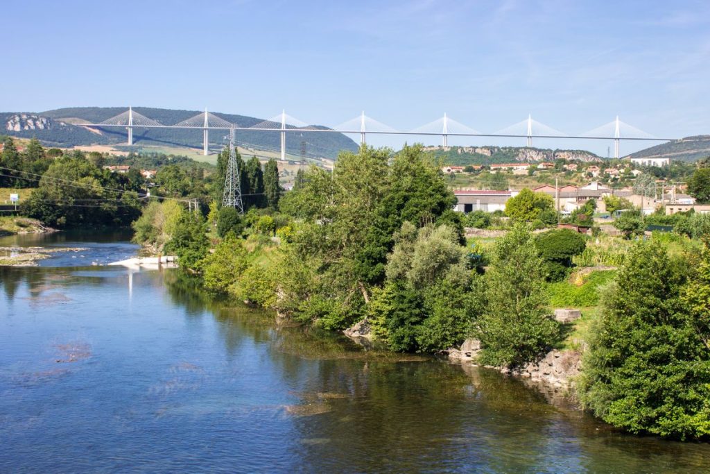 Une vue panoramique sur une rivière avec une verdure luxuriante au premier plan et un pont suspendu moderne en arrière-plan sous un ciel bleu clair.