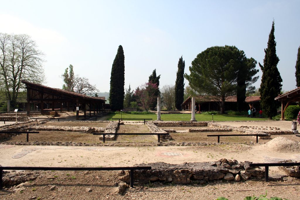 Ruines archéologiques aux contours de fondations visibles, entourées d'arbres et de pavillons couverts, sous un ciel dégagé.