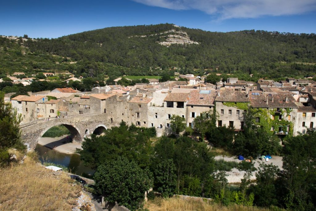 Un pont de pierre historique menant à un village traditionnel niché dans une vallée luxuriante avec des collines en arrière-plan.