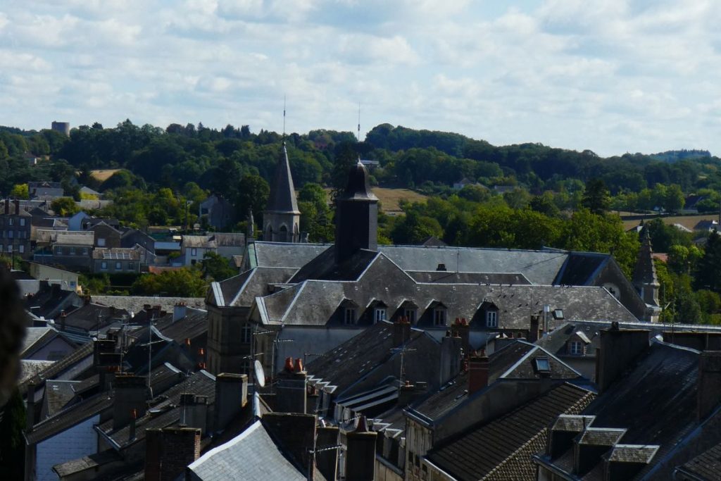 Une vue panoramique d'une ville européenne avec des bâtiments historiques et des clochers d'église sur fond de collines verdoyantes et un ciel partiellement nuageux.