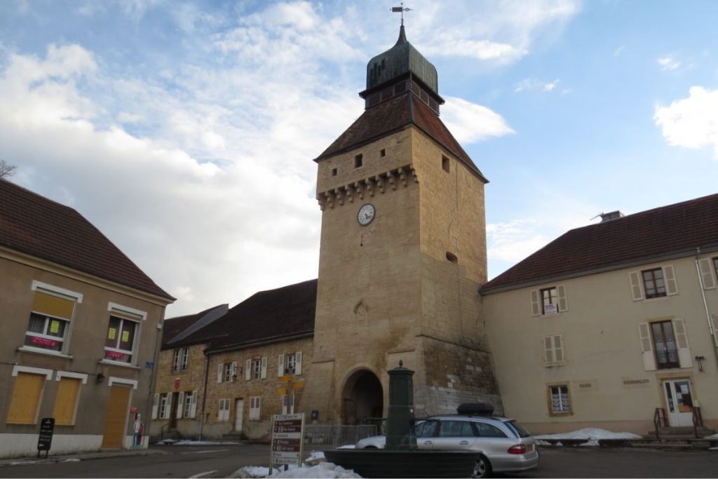 Une tour en pierre historique avec une horloge et une flèche, flanquée de bâtiments, sous un ciel nuageux. des plaques de neige sont visibles au sol. Une tour en pierre historique avec une horloge et une flèche, flanquée de bâtiments, sous un ciel nuageux. des plaques de neige sont visibles au sol.