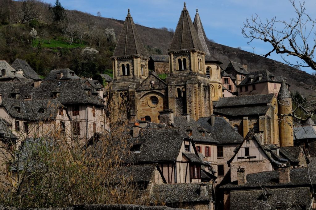 Village historique avec des bâtiments en pierre, des toits gris et une église à deux tours, niché à flanc de colline avec des arbres épars sous un ciel bleu clair. Village historique avec des bâtiments en pierre, des toits gris et une église à deux tours, niché à flanc de colline avec des arbres épars sous un ciel bleu clair.