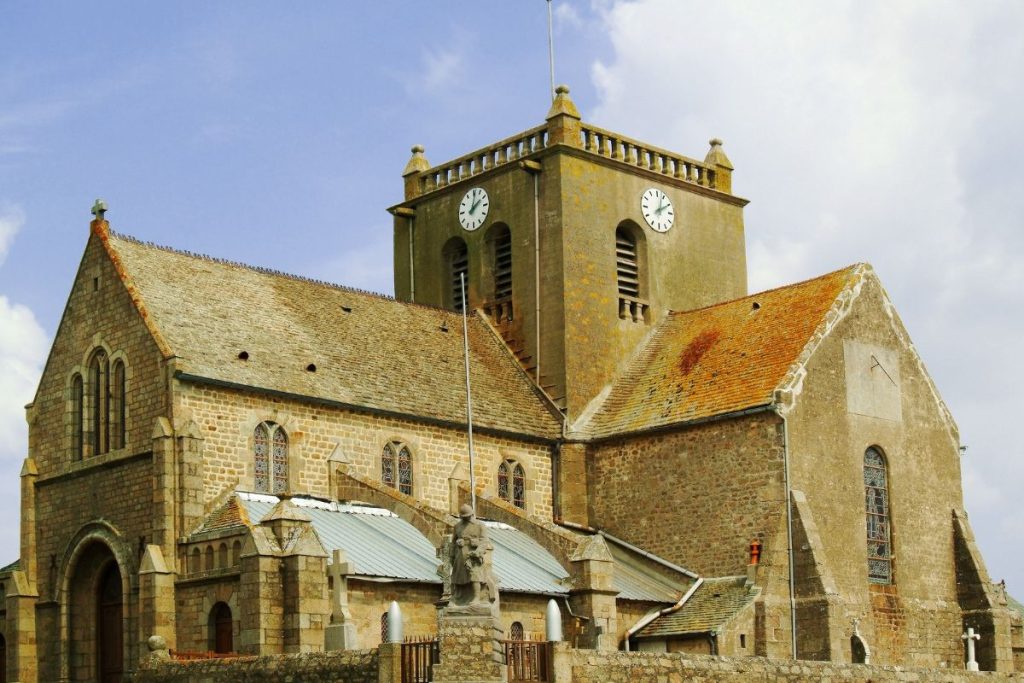 Une église en pierre avec une tour d'horloge, des fenêtres cintrées et une statue devant, se dresse sous un ciel partiellement nuageux. Une église en pierre avec une tour d'horloge, des fenêtres cintrées et une statue devant, se dresse sous un ciel partiellement nuageux.
