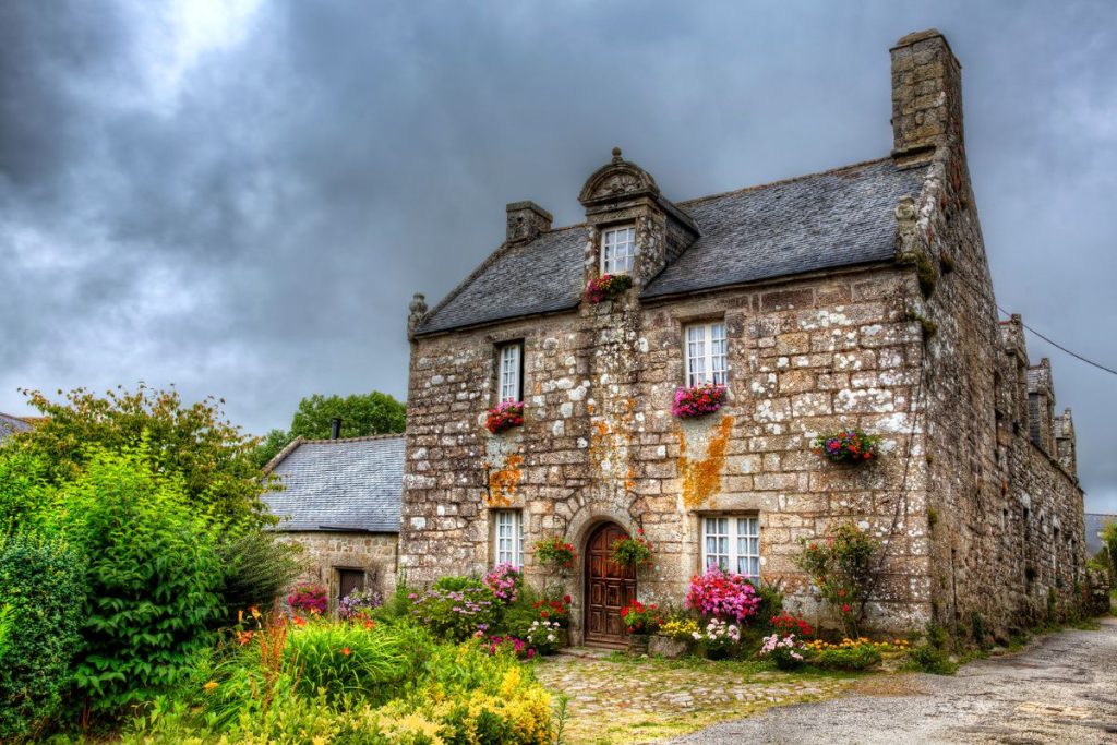 Une maison en pierre avec des bacs à fleurs et un jardin se dresse sous un ciel nuageux. Le bâtiment comprend une porte cintrée en bois, deux cheminées et de la verdure entourant l'entrée. Une maison en pierre avec des bacs à fleurs et un jardin se dresse sous un ciel nuageux. Le bâtiment comprend une porte cintrée en bois, deux cheminées et de la verdure entourant l'entrée.
