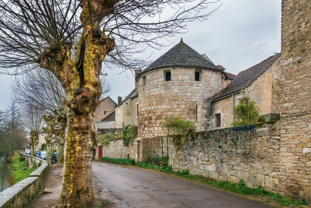 Une rue étroite bordée d'arbres sans feuilles et de bâtiments en pierre, dont une tour cylindrique en pierre, dans un village européen par temps couvert. Une rue étroite bordée d'arbres sans feuilles et de bâtiments en pierre, dont une tour cylindrique en pierre, dans un village européen par temps couvert.