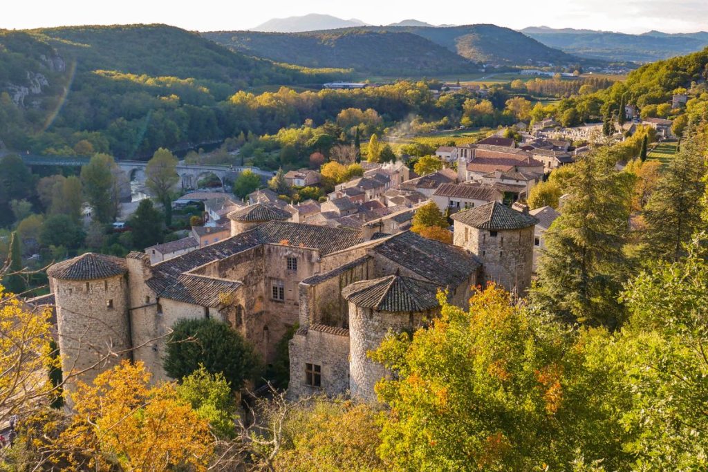 Vue aérienne d'un village historique avec des bâtiments en pierre et des tourelles entourées de verdure, avec des montagnes en arrière-plan. Vue aérienne d'un village historique avec des bâtiments en pierre et des tourelles entourées de verdure, avec des montagnes en arrière-plan.