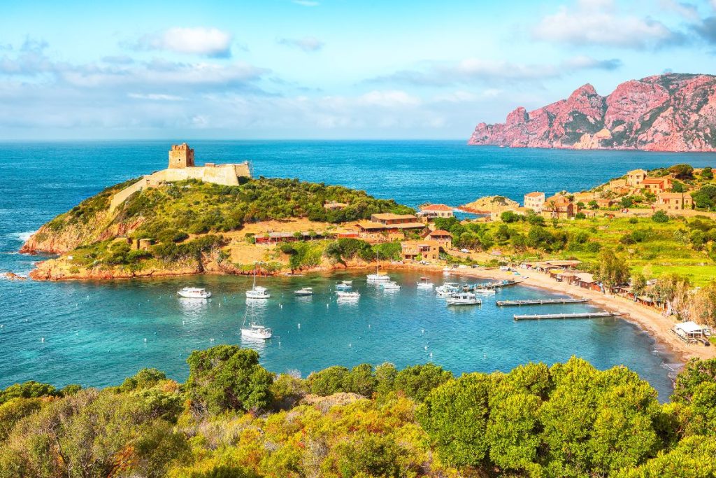 Paysage côtier avec une vieille forteresse sur une colline, des bateaux ancrés dans une petite baie, des bâtiments dispersés, une végétation luxuriante et des falaises rouges au loin, sous un ciel partiellement nuageux.